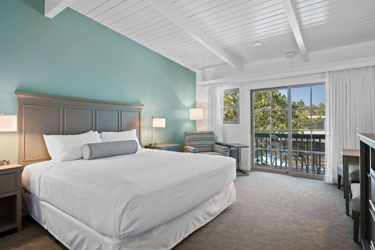 Une chambre d’hôtel avec un grand lit, une table de chevet avec une lampe, une chaise, une petite table et une vue sur un balcon donnant sur une piscine.