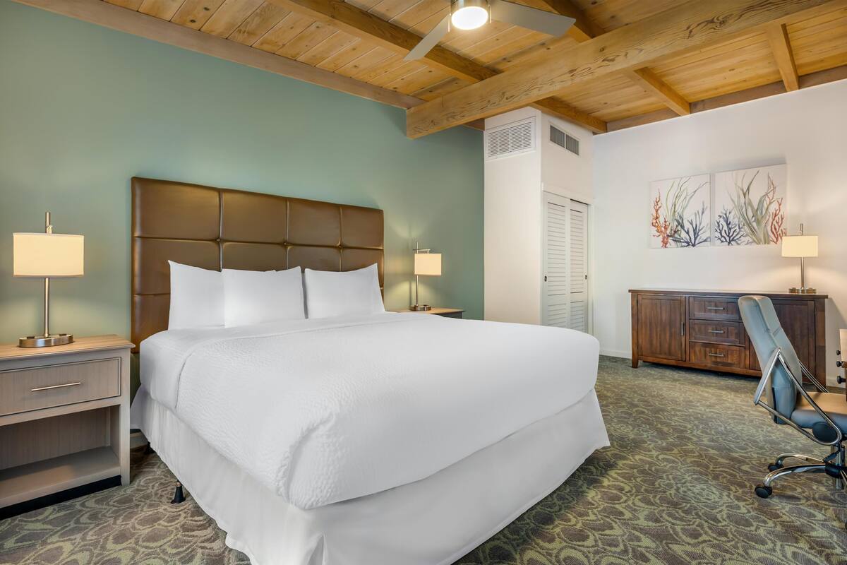 Une chambre d’hôtel avec un grand lit, deux lampes de chevet, une tête de lit en bois, un bureau avec une chaise et un mur décoré d’œuvres d’art.