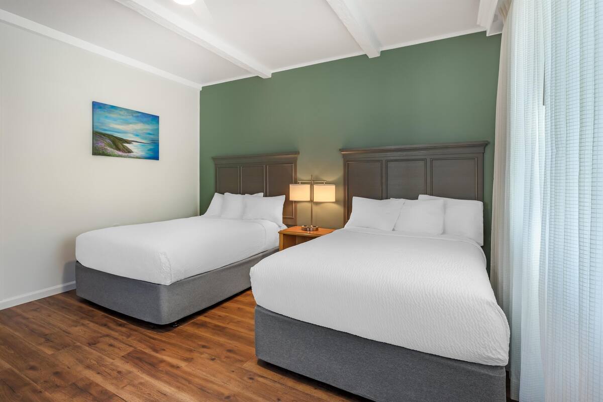 Une chambre d’hôtel avec deux lits simples, une table de chevet avec une lampe, un tableau au mur et un plancher en bois.