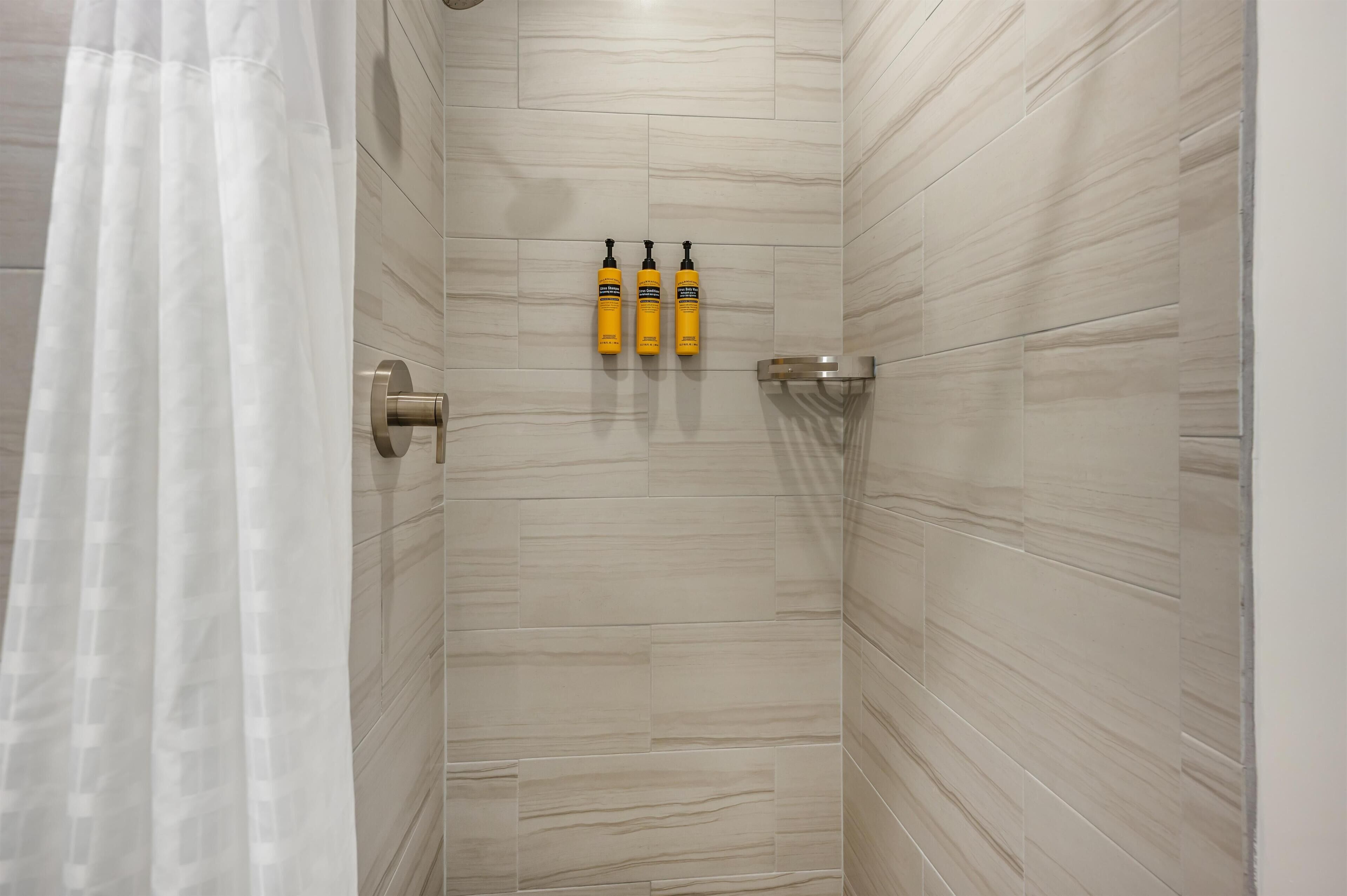 Una zona de ducha con azulejos color beige, una puerta de cristal y tres botellas amarillas en un estante.
