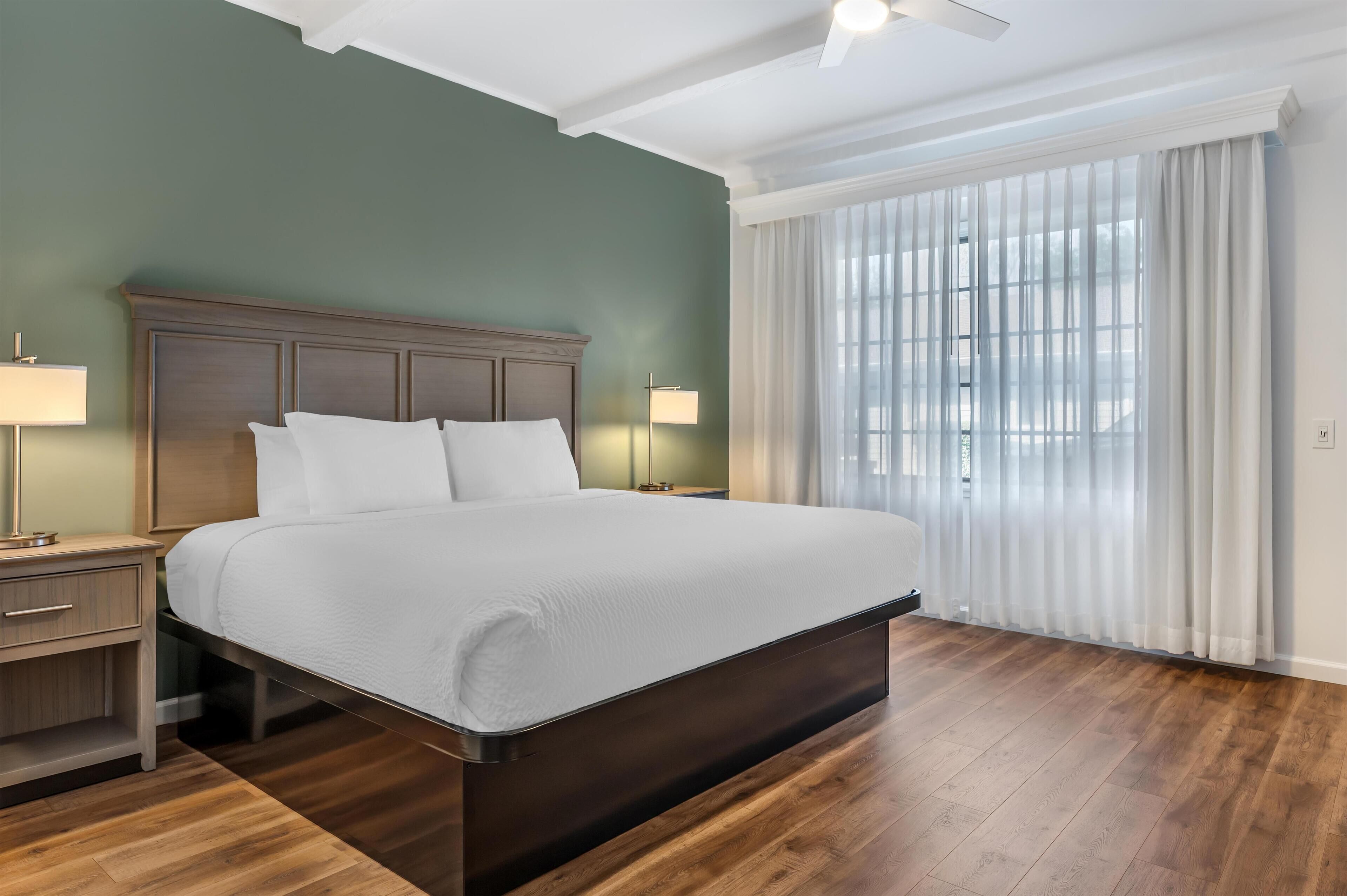 Una cama bien tendida en una habitación con piso de madera, pared verde y un ventanal con cortinas blancas.