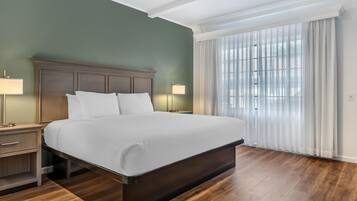 A neatly made bed in a room with a wooden floor, a green wall, and a large window with white curtains.