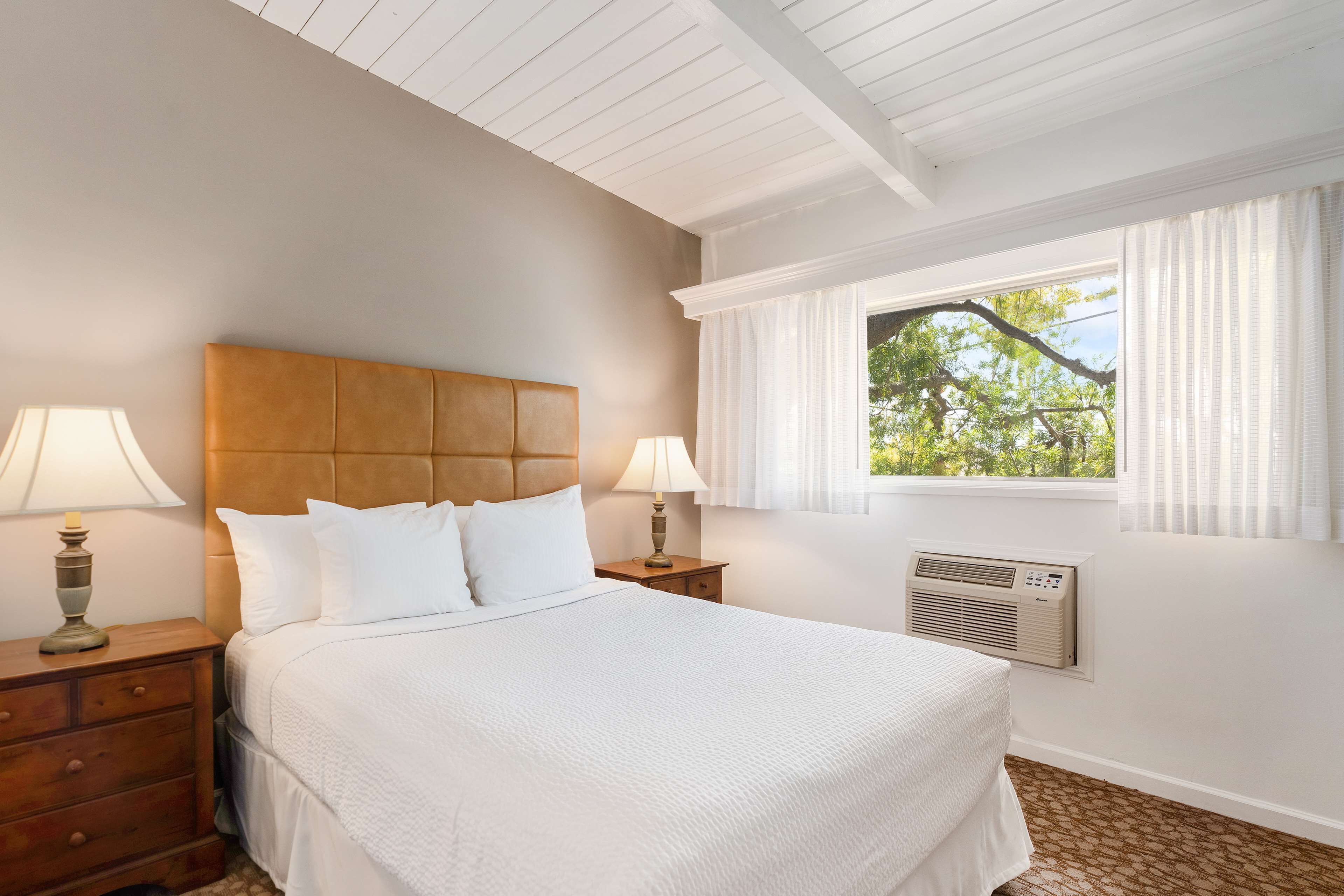 A bedroom with a large bed, two bedside tables with lamps, a window with curtains, and an air conditioning unit.