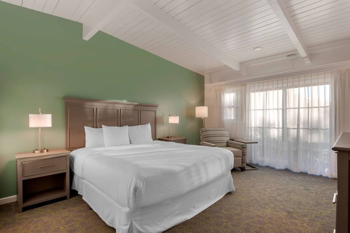 Une chambre d’hôtel avec un grand lit, deux tables de chevet avec des lampes, une chaise et une fenêtre avec des rideaux légers.