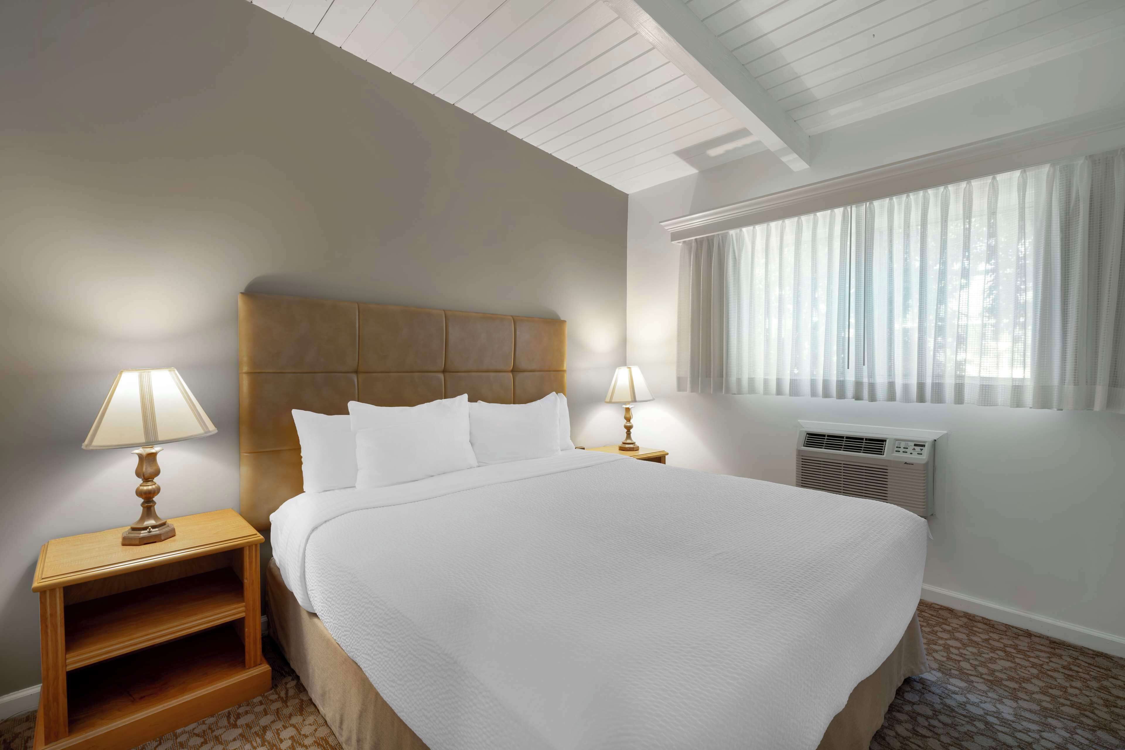 Ein Hotelzimmer mit einem großen Bett, zwei Nachttischlampen, einem Holznachttisch, einem Fenster mit Vorhängen und einer Klimaanlage.