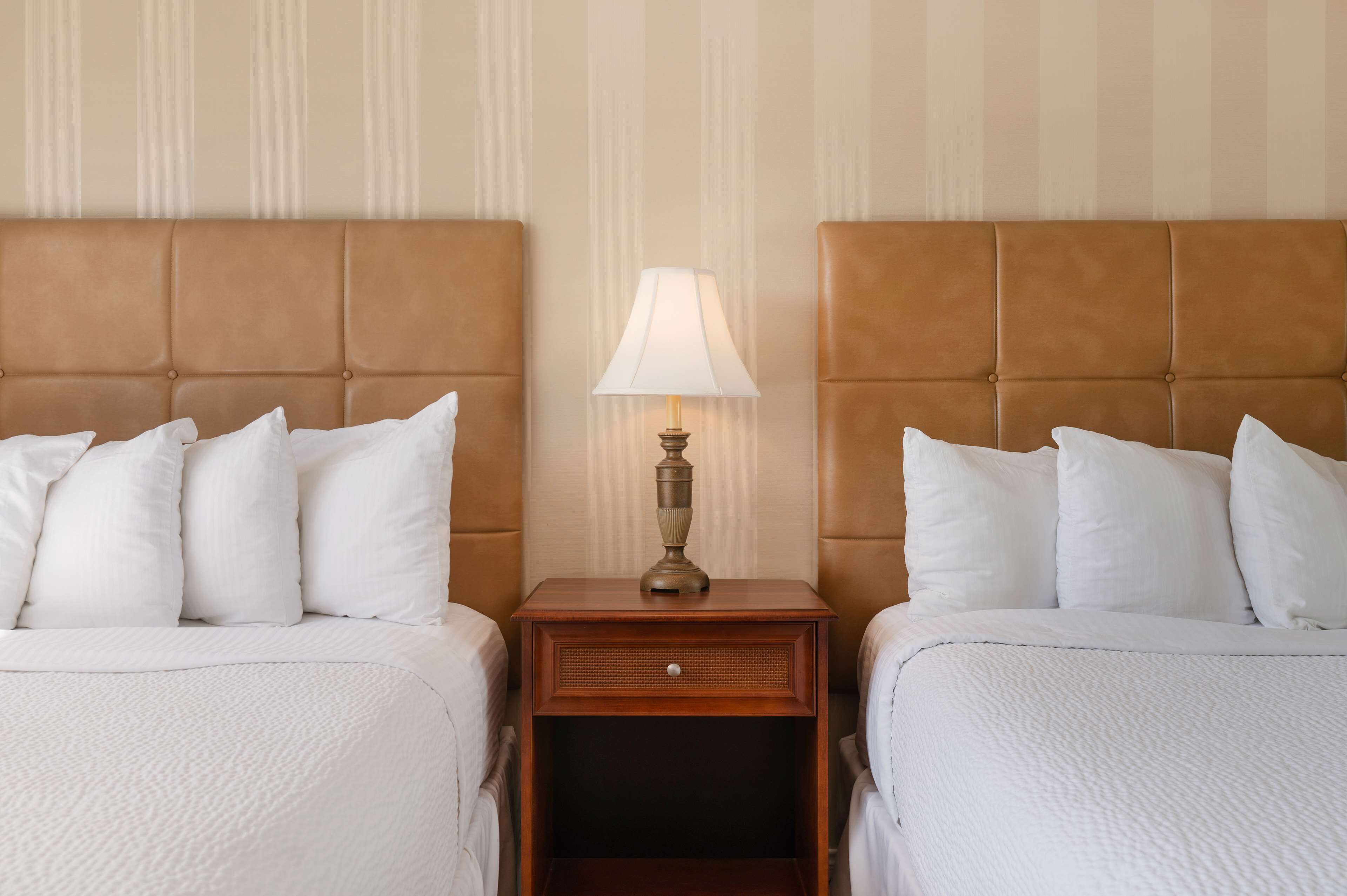 A hotel room with two beds, a nightstand with a lamp, and a striped wall.