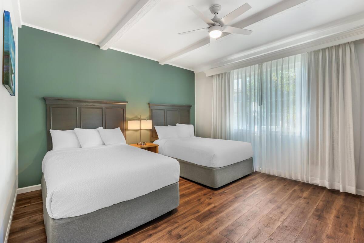 Une chambre d’hôtel avec deux lits, un ventilateur de plafond, une table de chevet avec une lampe et une grande fenêtre avec des rideaux légers.
