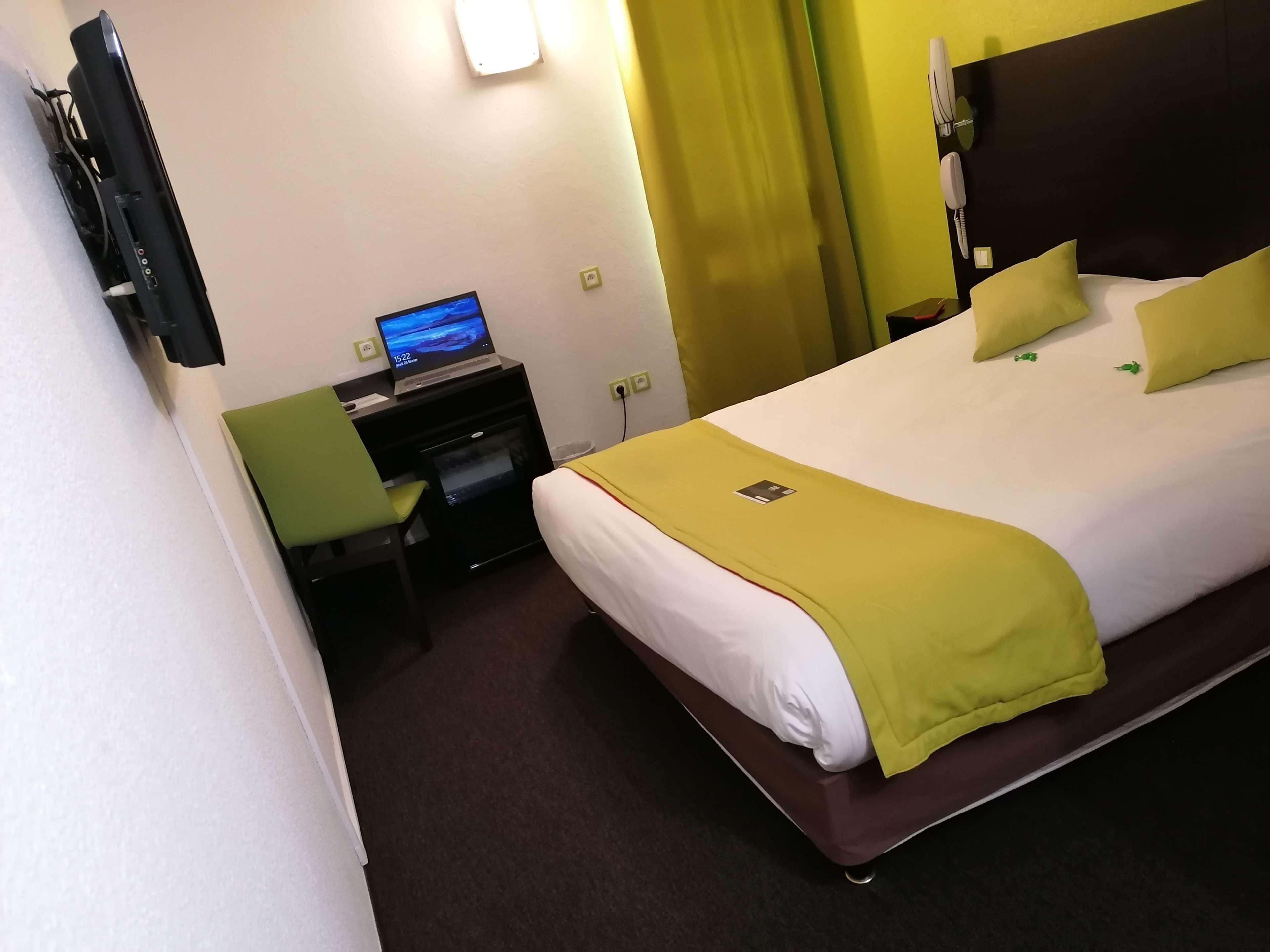 Ett hotellrum med en säng, ett skrivbord med en bärbar dator, en telefon och en TV som är monterad på väggen.