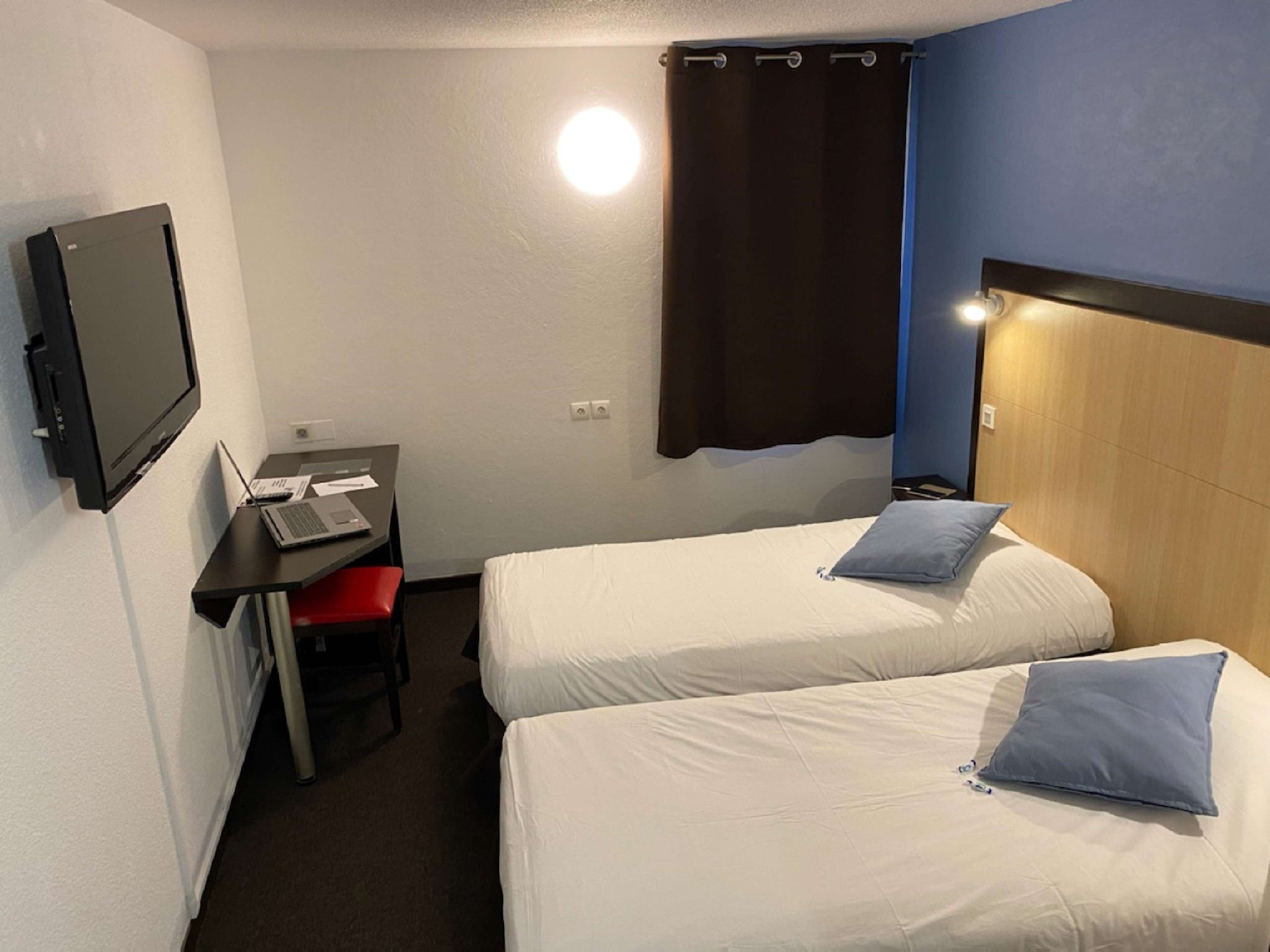 Ett hotellrum med två separata sängar, ett skrivbord med en bärbar dator, en tv som är monterad på väggen och en gardin.