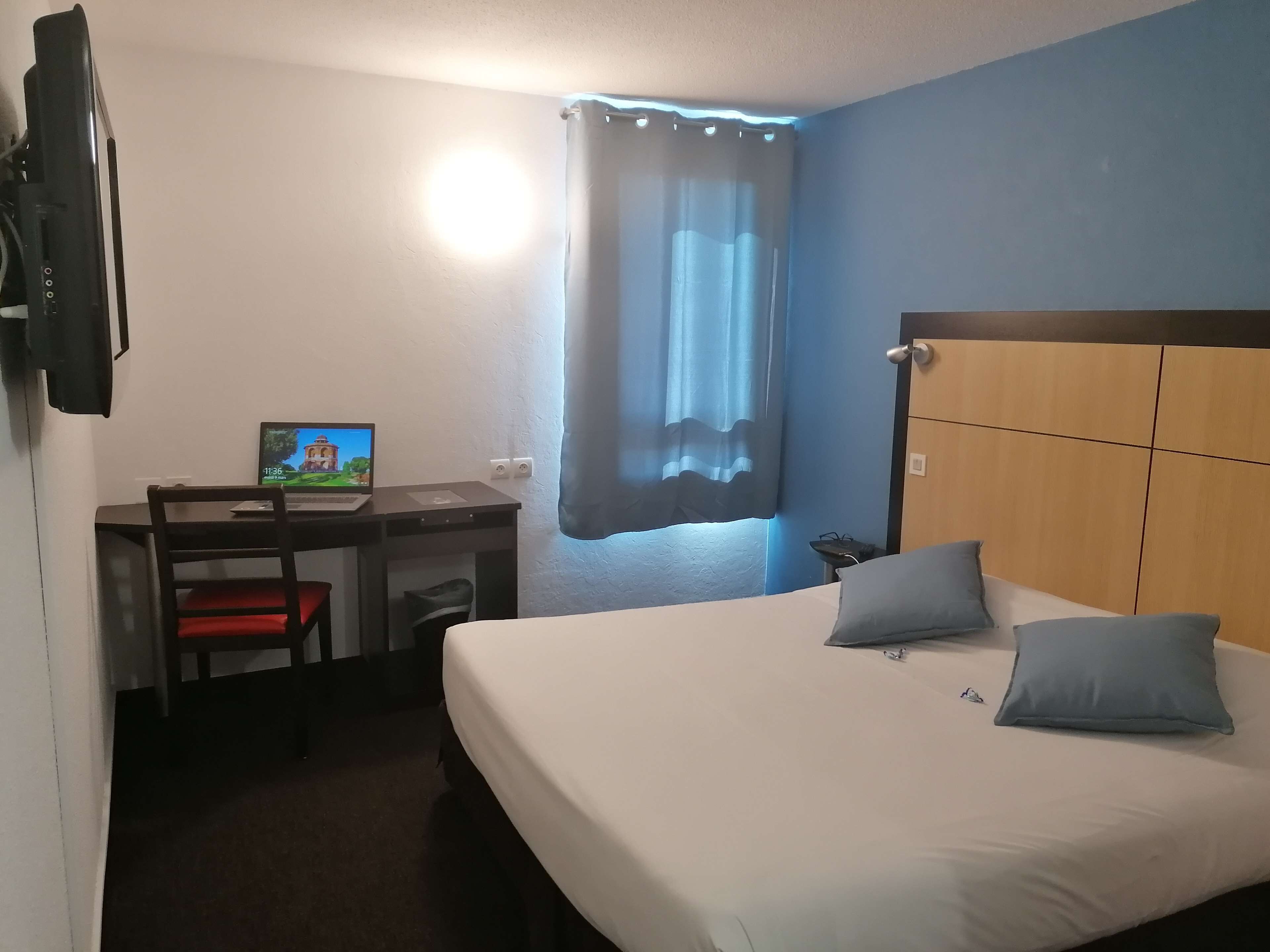 A hotel room with a bed, a desk with a laptop, a chair, a TV, and a window with curtains.