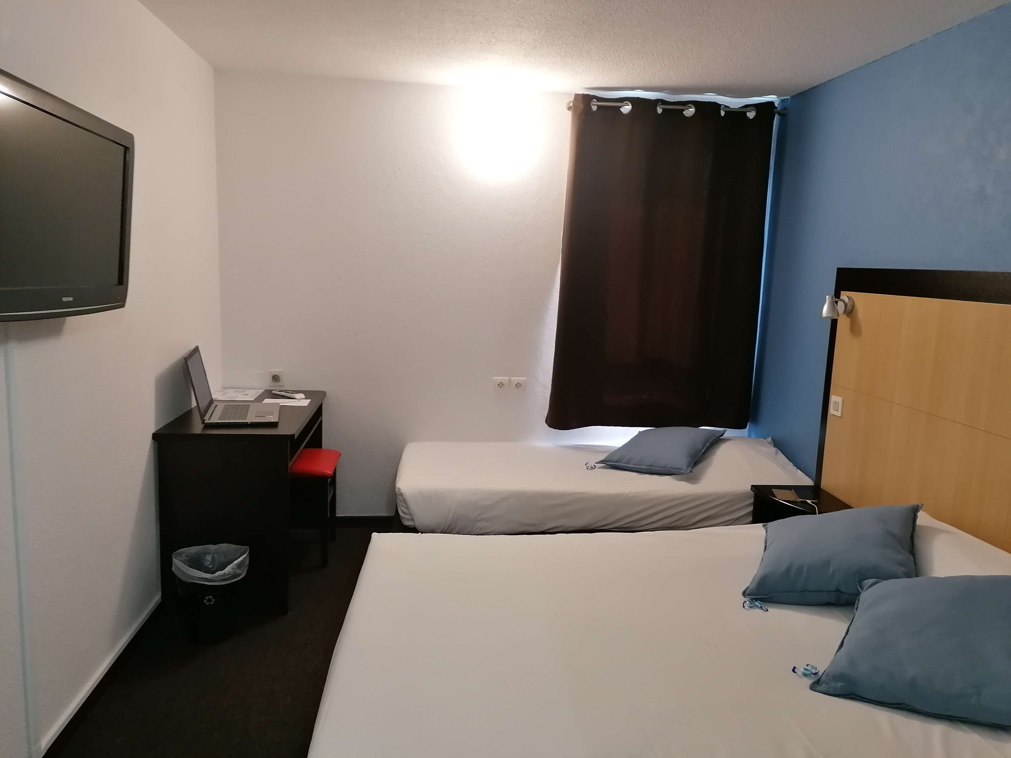 Quarto de hotel com duas camas, uma mesa com um laptop, uma televisão e uma janela com cortinas.