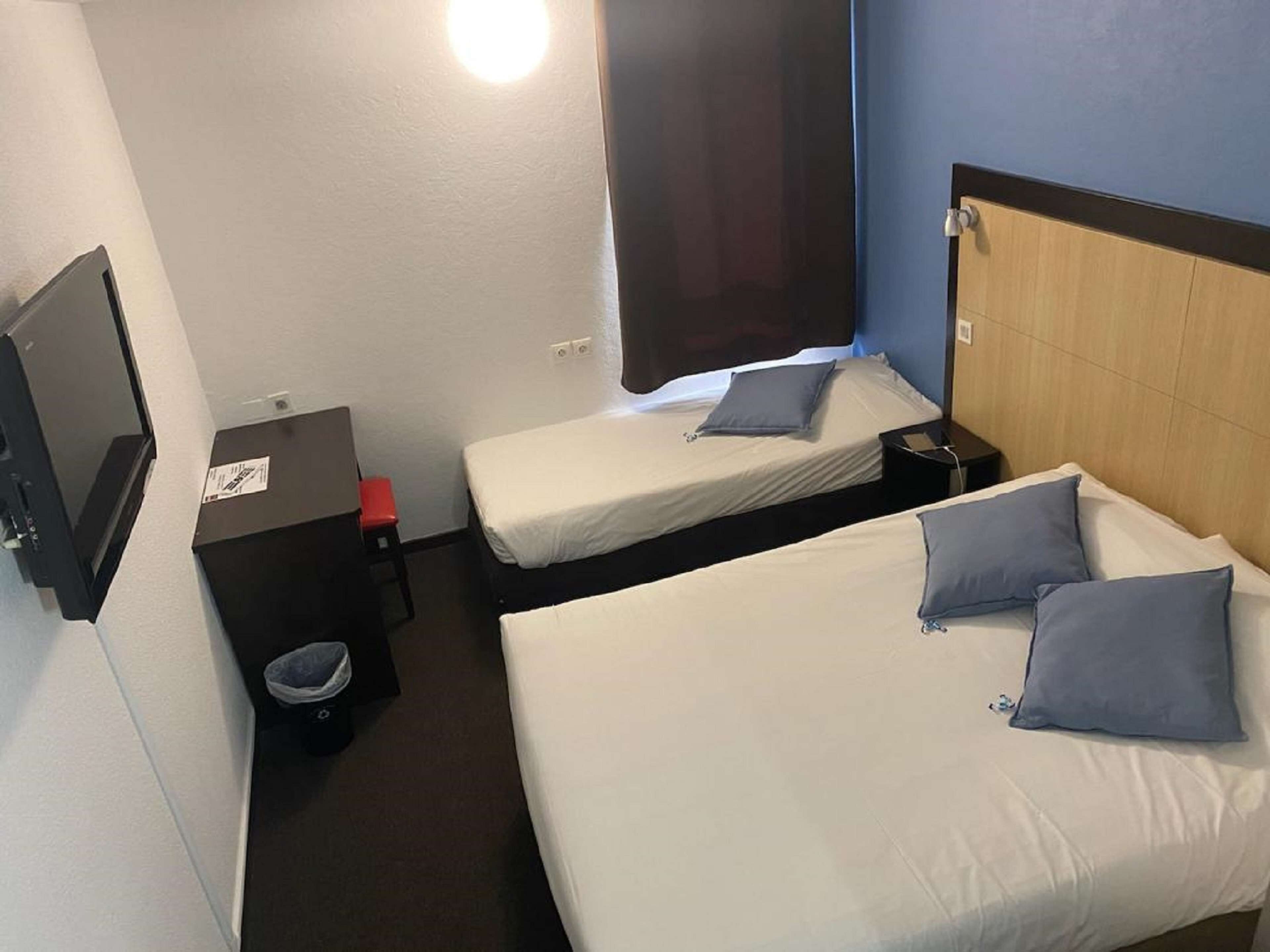 Ein Hotelzimmer mit zwei Einzelbetten, einem kleinen Schreibtisch mit Stuhl, einer Mülleimer und einem an der Wand befestigten Fernseher.