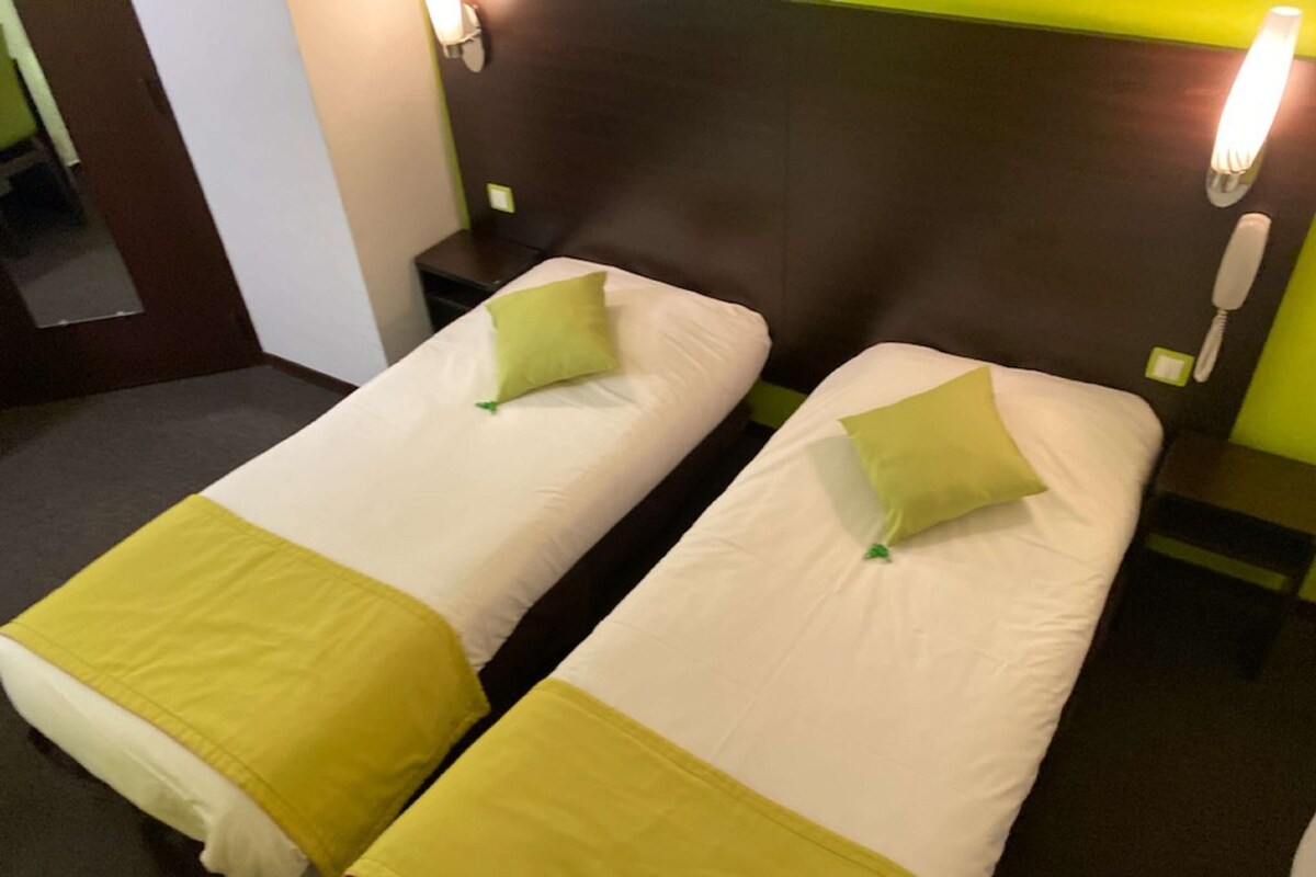 A hotel room with two single beds, each with a green pillow and a yellow bedspread. There are bedside lamps and a phone on the nightstand.