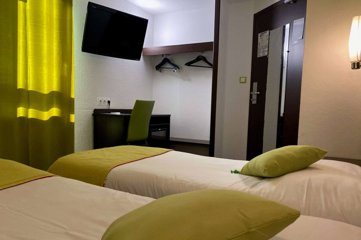 Ein Hotelzimmer mit zwei Betten, jedes mit einem grünen Kissen, einem Schreibtisch mit Stuhl, einem an der Wand montierten Fernseher und einer Tür mit Kleiderhaken.