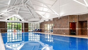An indoor swimming pool with clear glass walls, a tiled floor, and a ceiling with white beams. - Macdonald Frimley Hall Hotel and Spa (Camberley)
