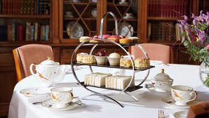 A three-tiered tea service stand with sandwiches and pastries, surrounded by tea cups and saucers on a table. - Macdonald Frimley Hall Hotel and Spa (Camberley)