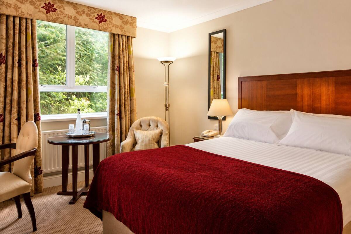 Habitación de hotel con cama, una silla, una mesita, una lámpara y ventana con cortinas.