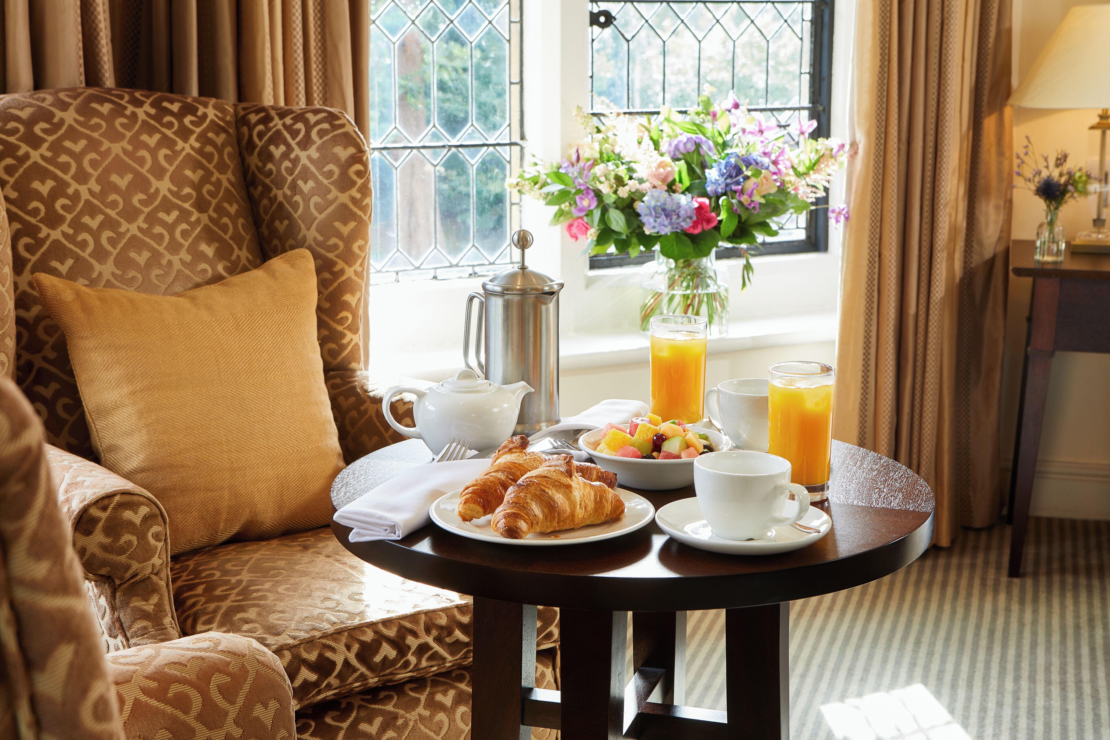 A breakfast setup with croissants, fruit, juice, and a coffee cup on a table, with a patterned armchair and a window with curtains in the background.