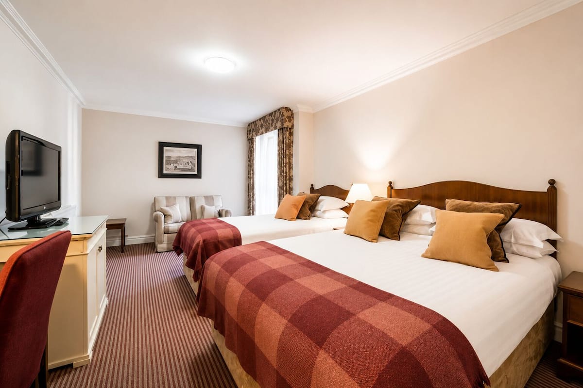 Habitación de hotel con dos camas, un escritorio con televisor, una silla, un cuadro en la pared y una ventana con cortinas.