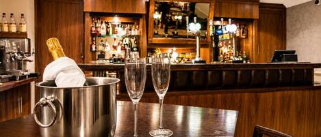 Zona de bar con un cubo metálico que contiene una botella de champán, dos copas de vino vacías y un mostrador de bar de madera con varias botellas y una caja registradora.