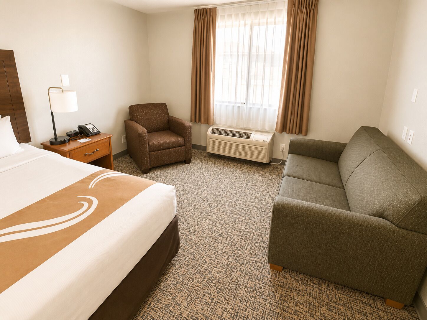 Una habitación de hotel con una cama, una silla, un sofá, un escritorio con teléfono y un equipo de aire acondicionado.