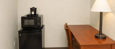 A small kitchenette with a microwave and a refrigerator, a wooden dining table with a chair, and a floor lamp.