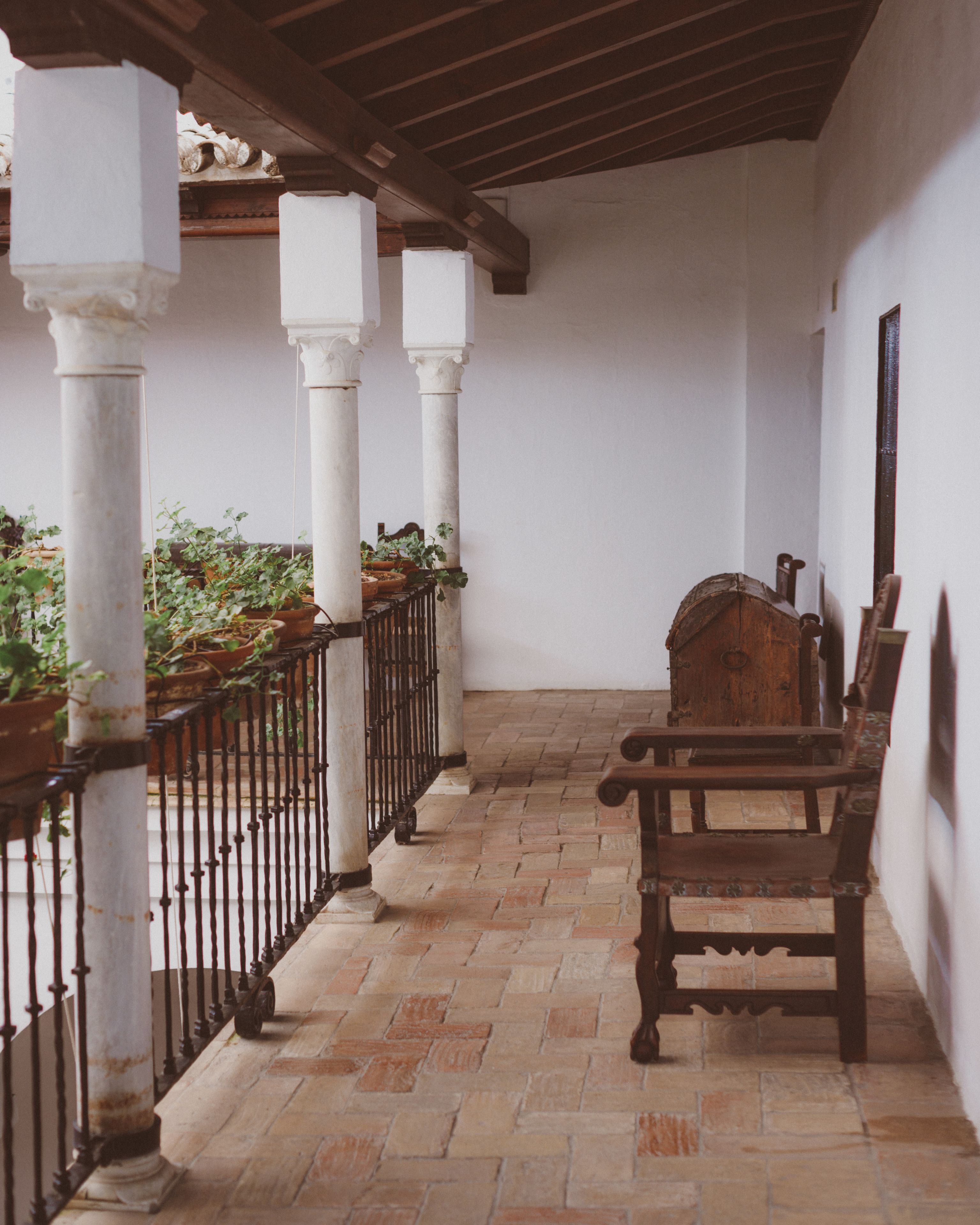 Un espacio al aire libre techado con un banco de madera, plantas en macetas y piso de baldosas.