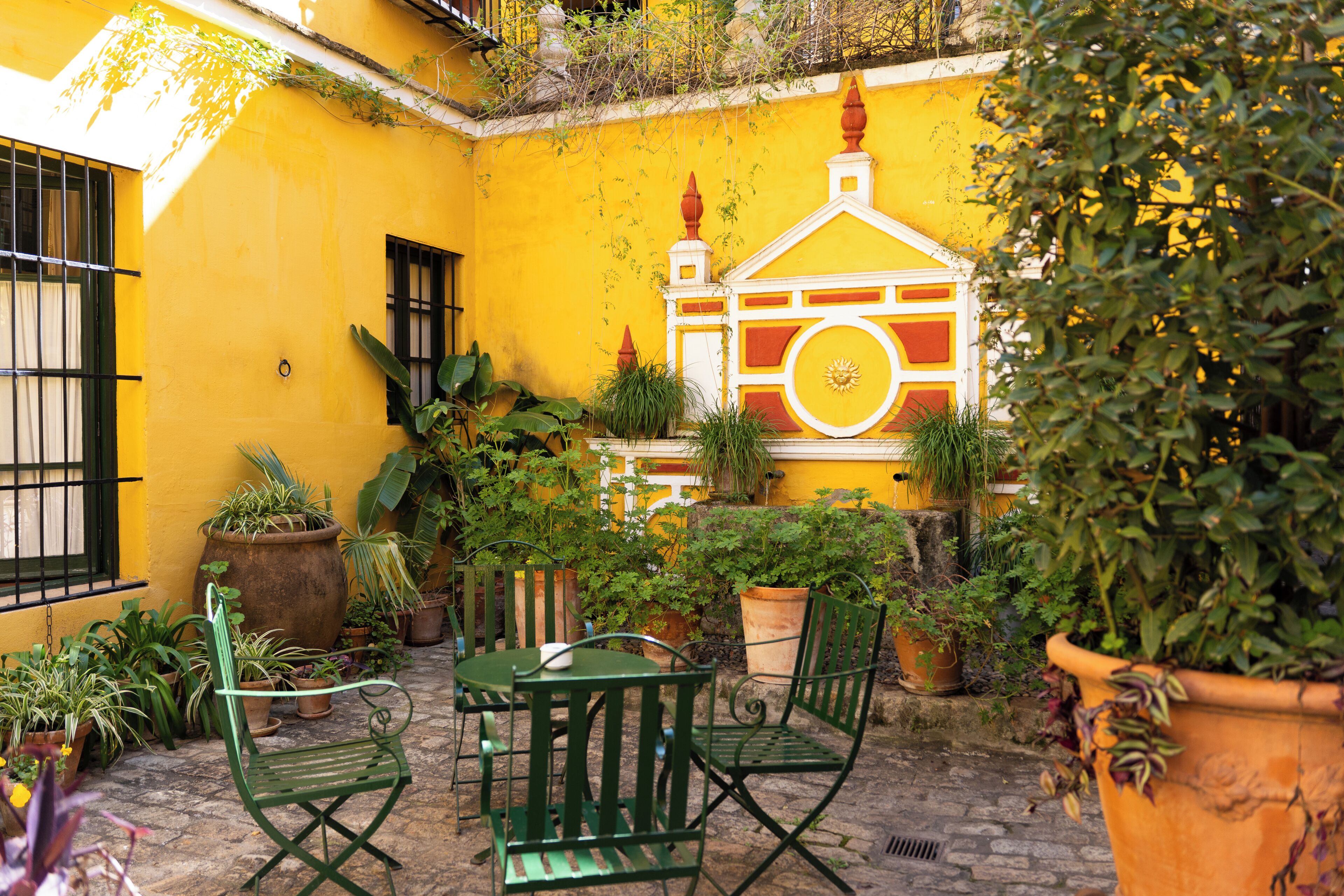 Une cour avec des murs jaunes, des chaises en métal vertes, une table et des plantes en pot.