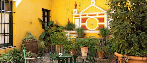 Un patio con paredes amarillas, sillas de metal verde, una mesa y plantas en macetas.