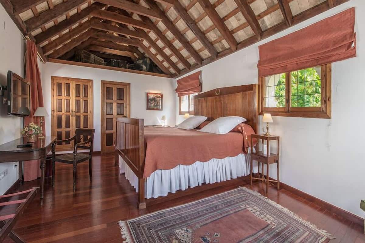 A bedroom with a wooden ceiling, a bed with a white and pink bedspread, a desk with a chair, and a window with a view of greenery.