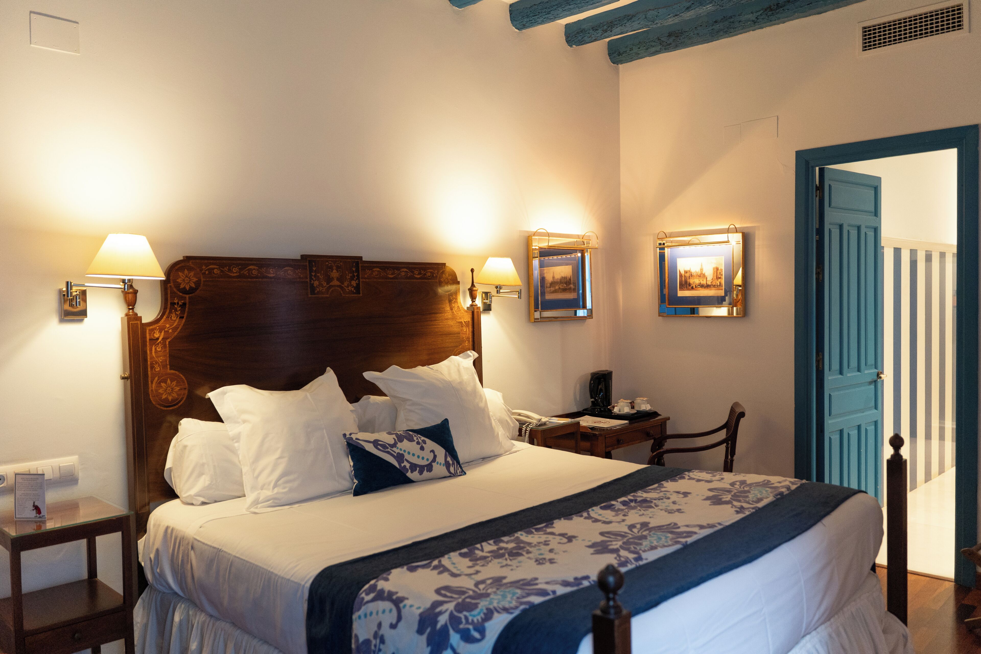 A neatly made bed with a wooden headboard, two bedside tables with lamps, a small desk, and a door with a blue frame.