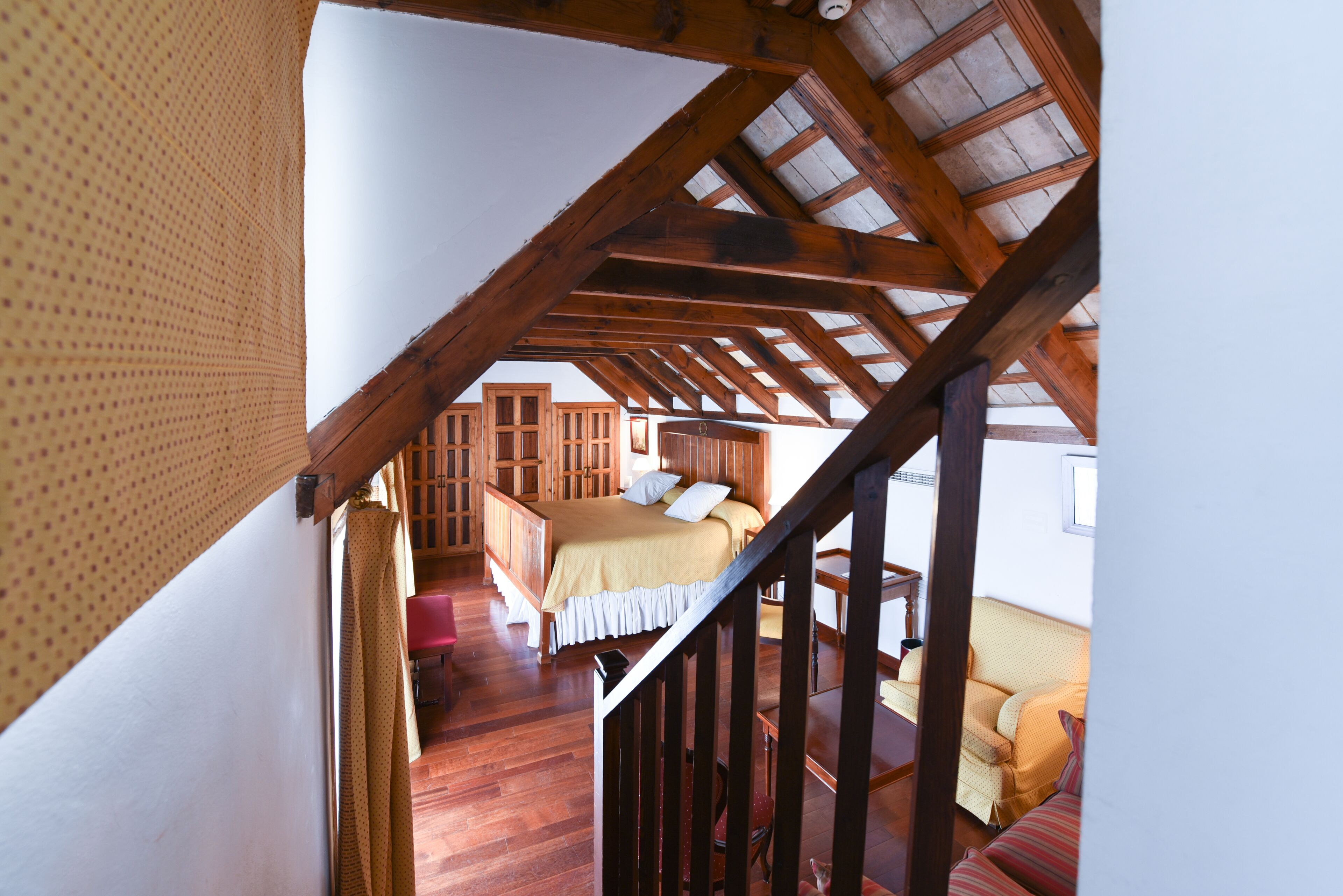 Une chambre avec un plafond en bois, un lit recouvert d’une courtepointe blanche et beige, et une commode en bois.