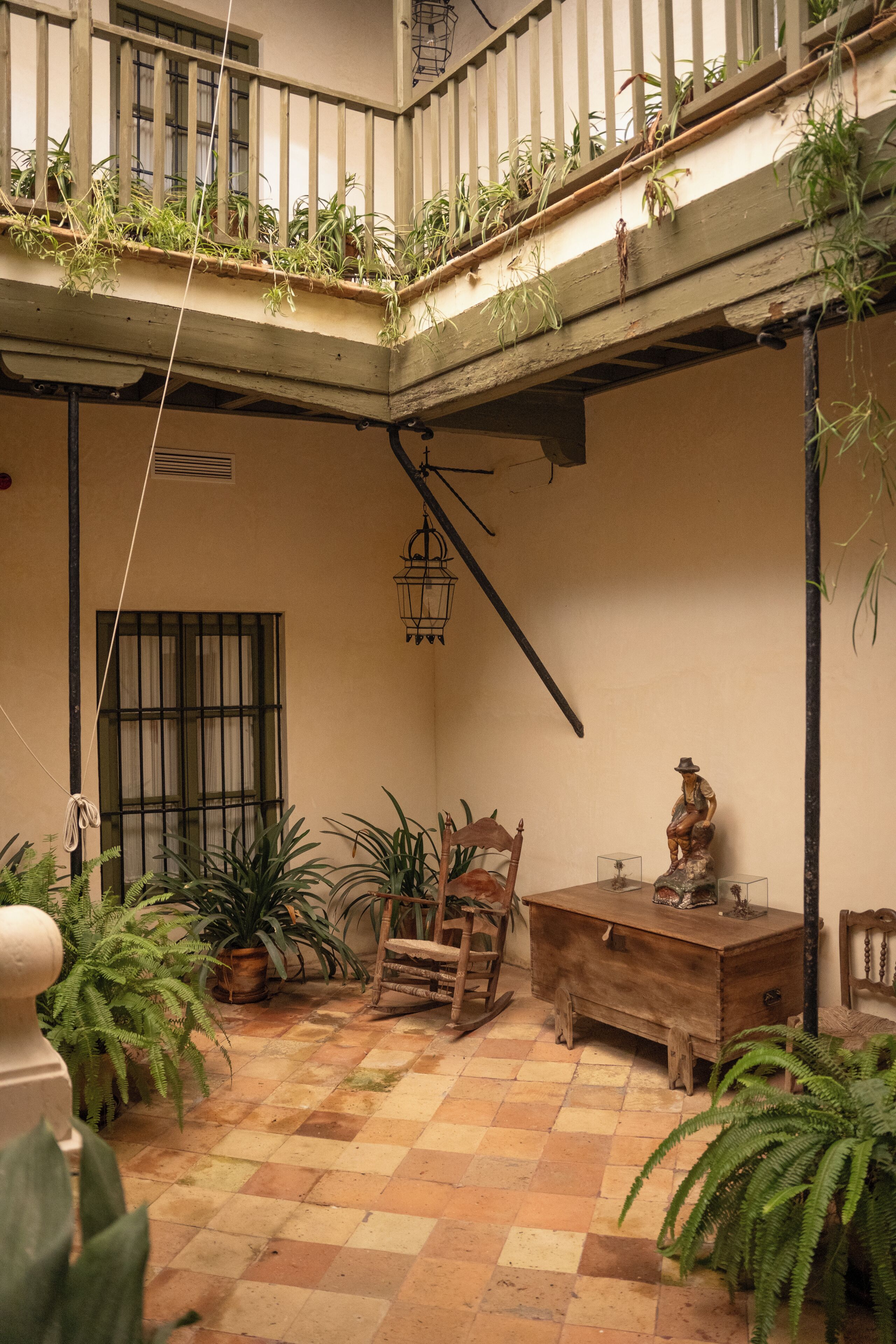 Un patio con suelo a cuadros, mobiliario de madera, plantas en macetas y un balcón con barandillas.