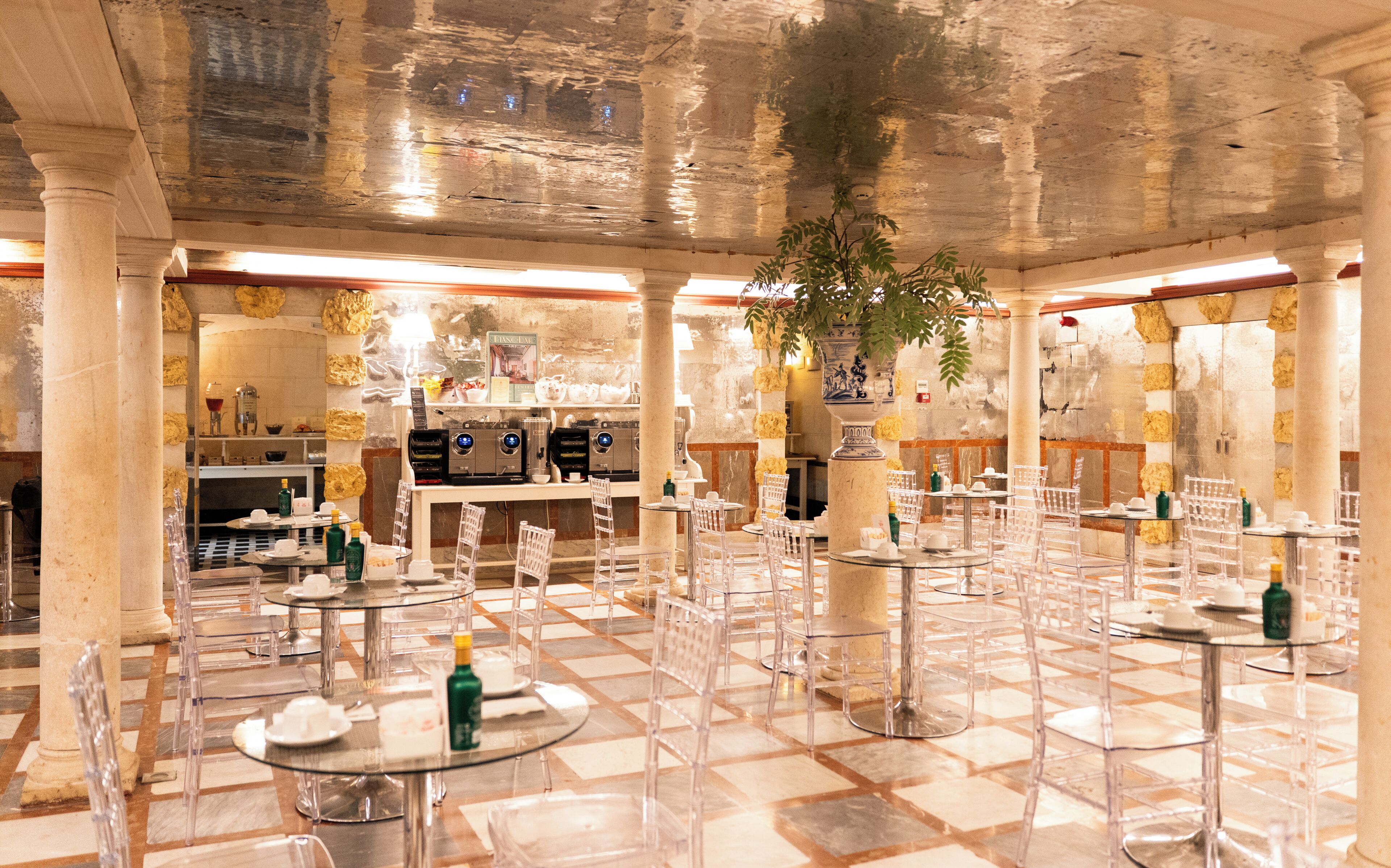 A restaurant with marble flooring, clear chairs, and tables, featuring a bar area in the background.