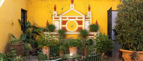 A courtyard with yellow walls, a decorative gate, potted plants, and green chairs.