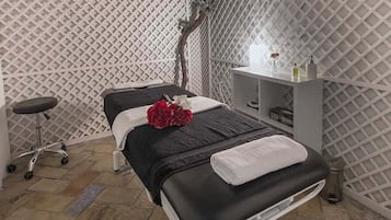 Una stanza per massaggi con un lettino da massaggio, uno sgabello, una mensola con flaconi e una pianta decorativa.