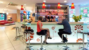 A bar area with a counter, stools, and a bartender. There are shelves with bottles behind the bar. - ibis Mulhouse Centre Filature (Mulhouse)