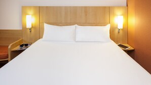 A hotel room with a wooden headboard, two bedside lamps, a white bed, a nightstand with a smartphone, and a magazine. - ibis Mulhouse Centre Filature (Mulhouse)