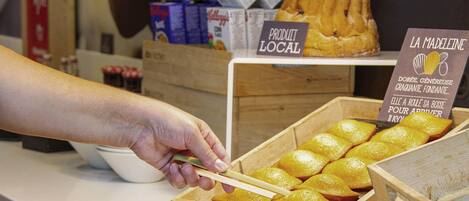 Una mano elige pasteles de una caja de madera, con cajas de cereal y un letrero que indica productos locales al fondo.