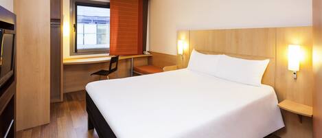 A hotel room with a large bed, a desk with a chair, and a window with blinds.