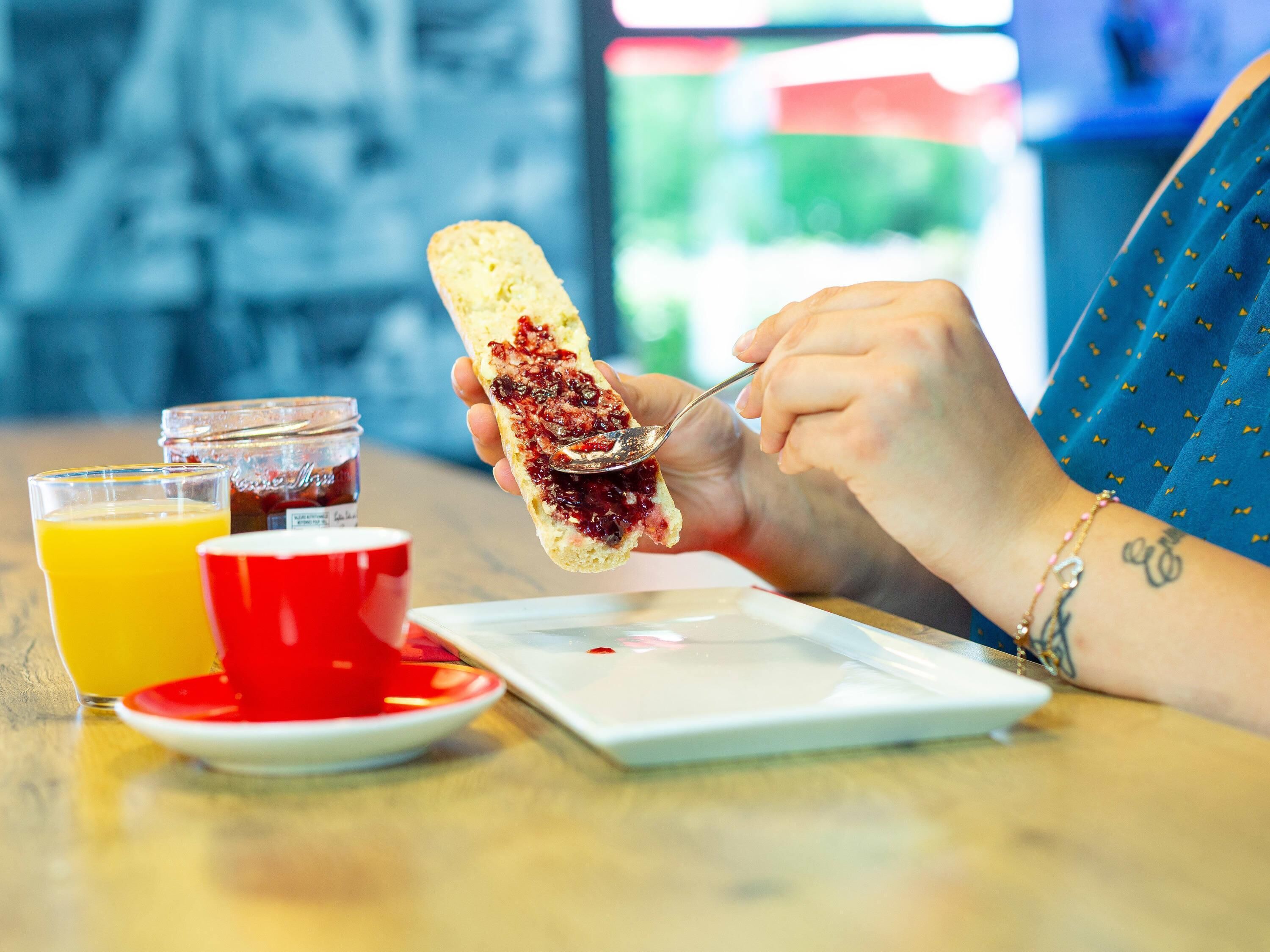 A person eating a sandwich with jam, a glass of orange juice, and a red cup on a table.
