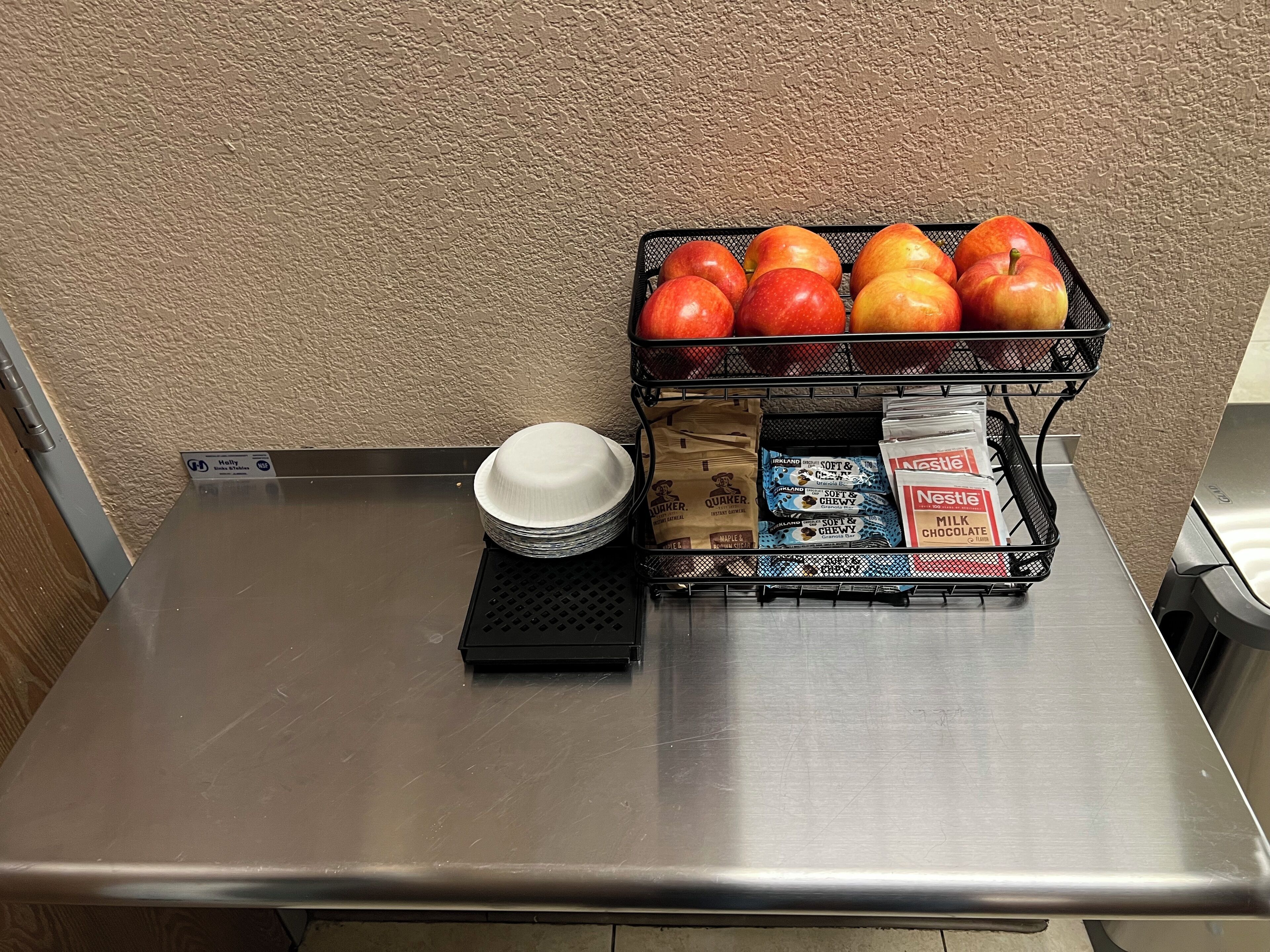 Una mesa de acero inoxidable con una pila de platos de papel blancos, una cesta negra con manzanas y una bandeja de barras de chocolate Nestle.