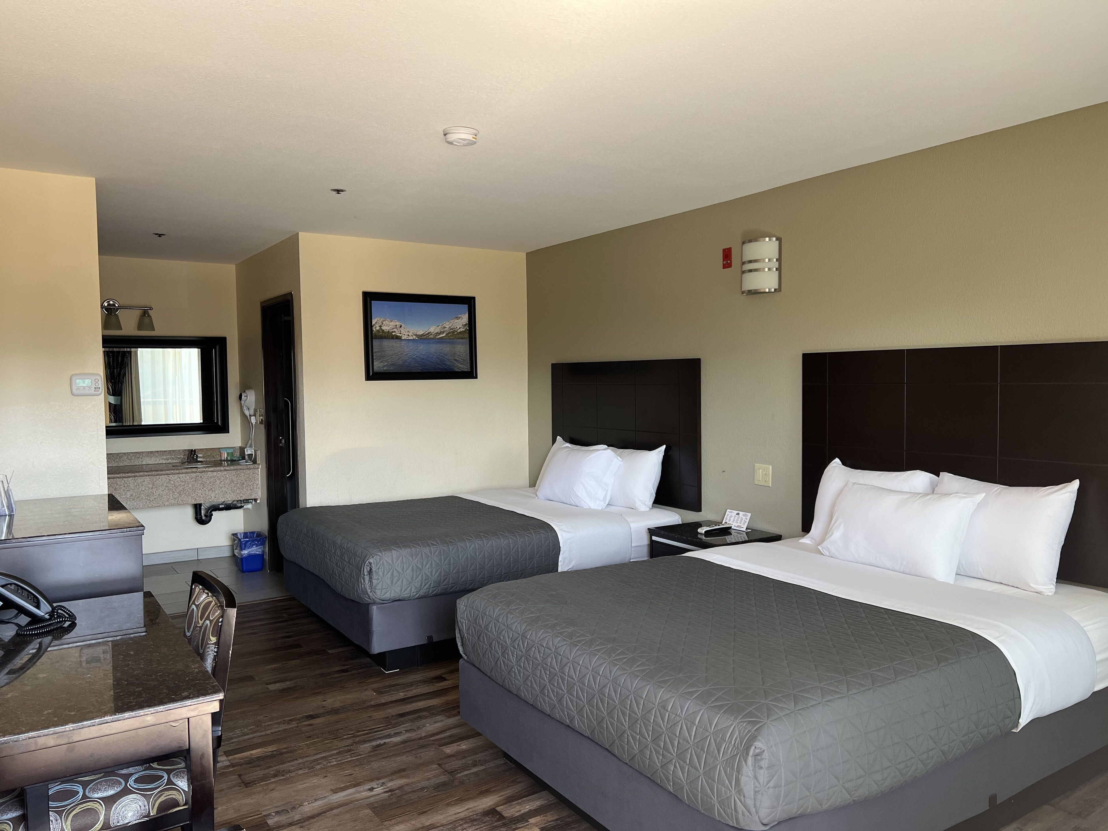 A hotel room with two beds, a desk, a mirror, and a painting of a lake.