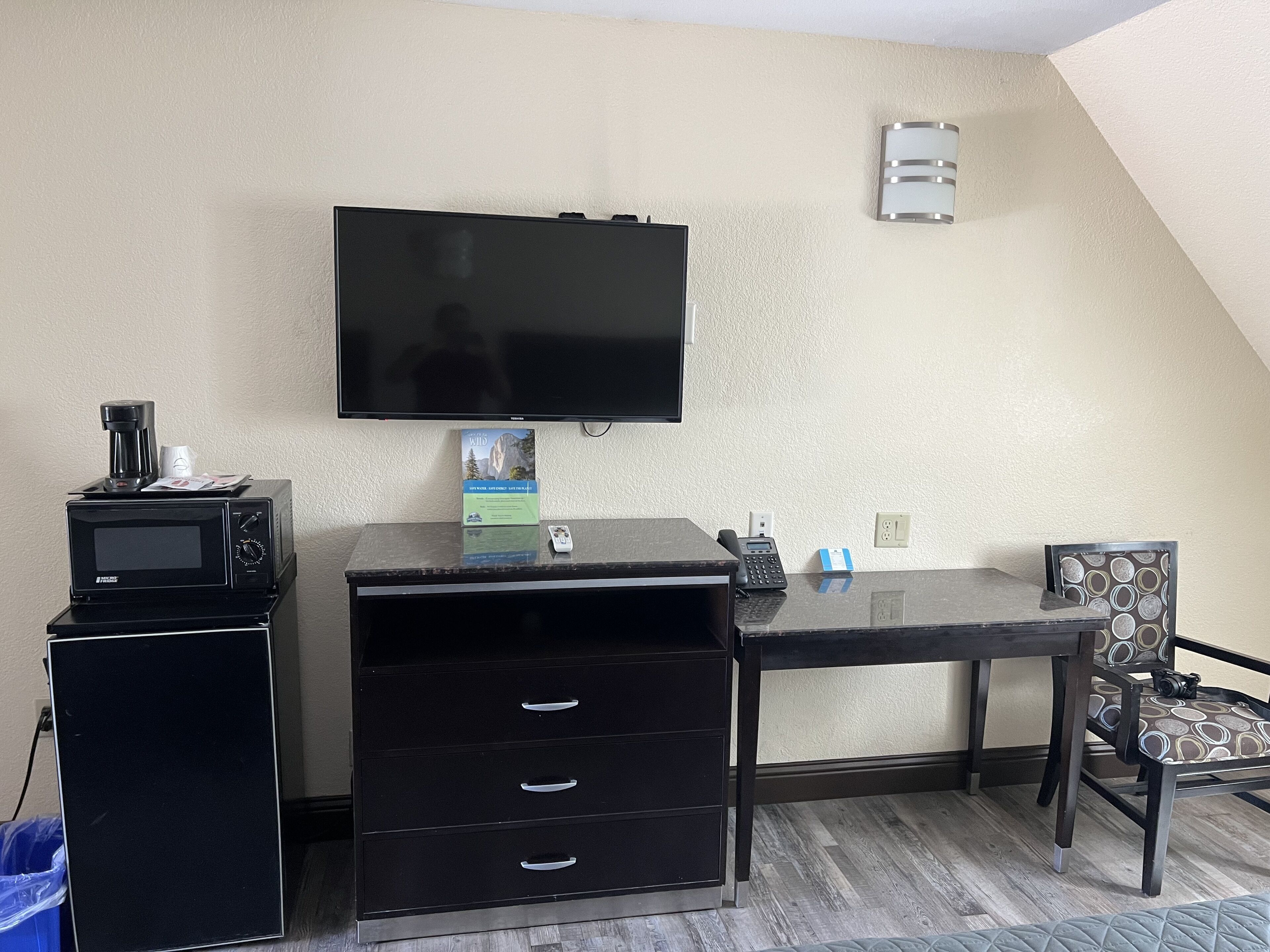 A compact hotel room with a flat-screen TV mounted on the wall, a small desk with a microwave, a coffee maker, and a book. There is a chair with a patterned cushion and a camera on it.