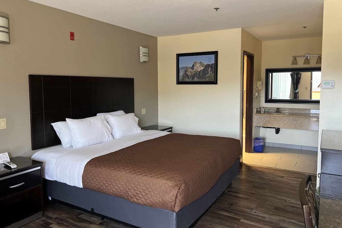 A hotel room with a bed, a nightstand, a wall-mounted lamp, a painting, a bathroom with a sink and mirror, and a wooden floor.