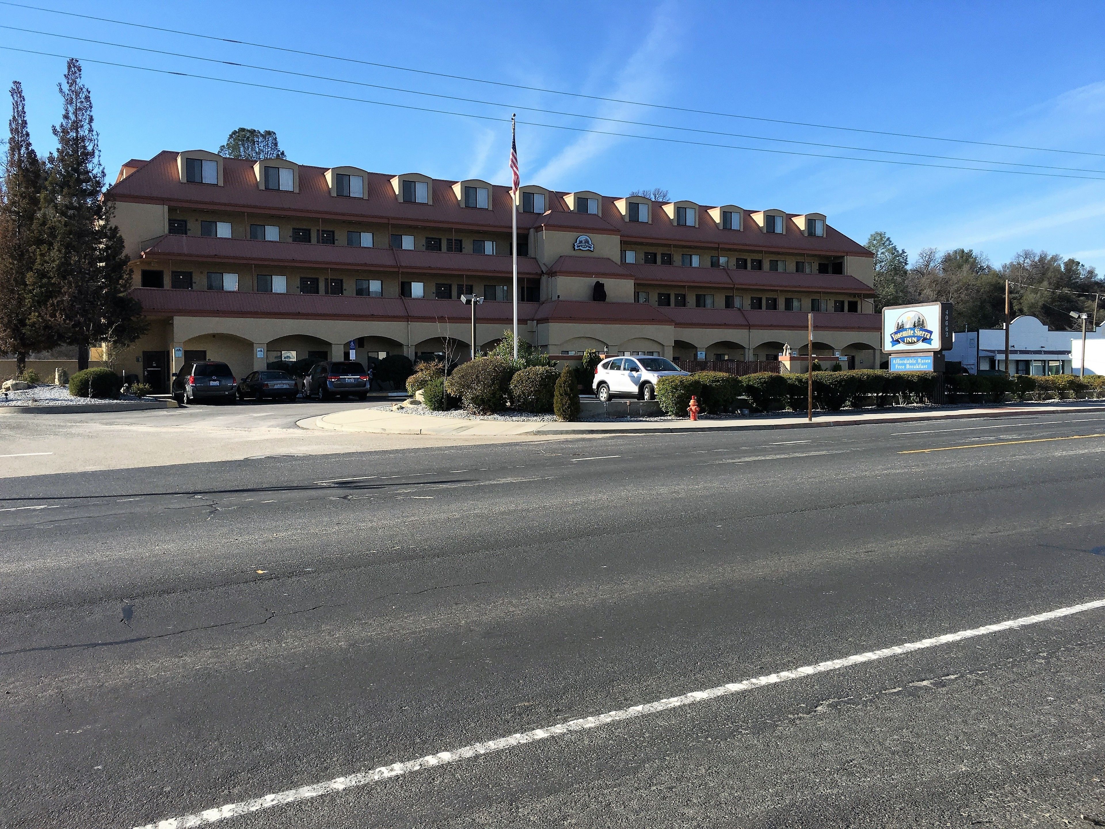 Un hotel de varios pisos con techo rojo y paredes color beige, que cuenta con un estacionamiento con varios autos y una bandera estadounidense visible.