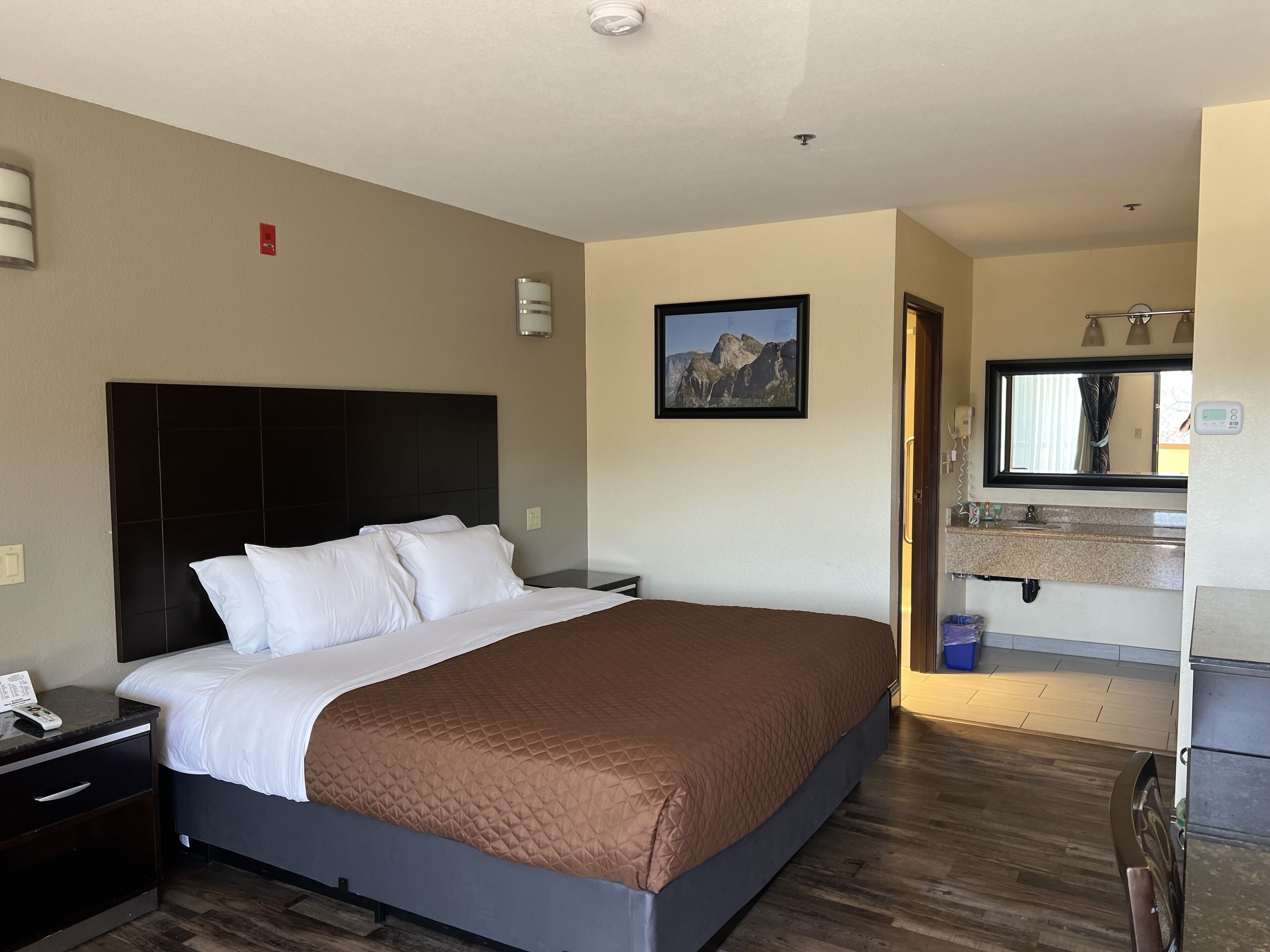 Habitación de hotel con una cama grande, una mesita de noche, un baño con lavabo y espejo, y una fotografía de montañas en la pared.