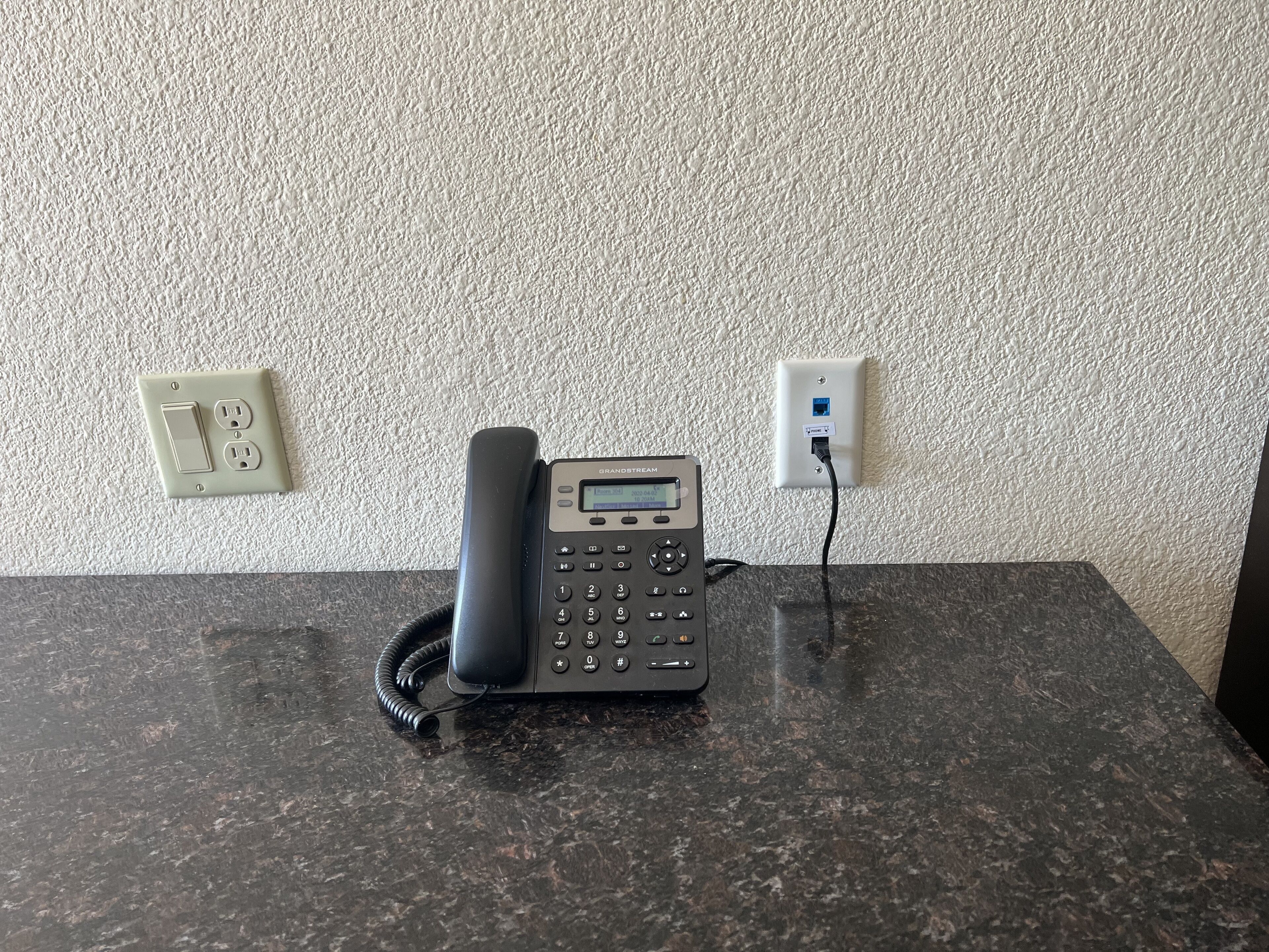 A black desk phone on a granite countertop with a textured wall and electrical outlets in the background.