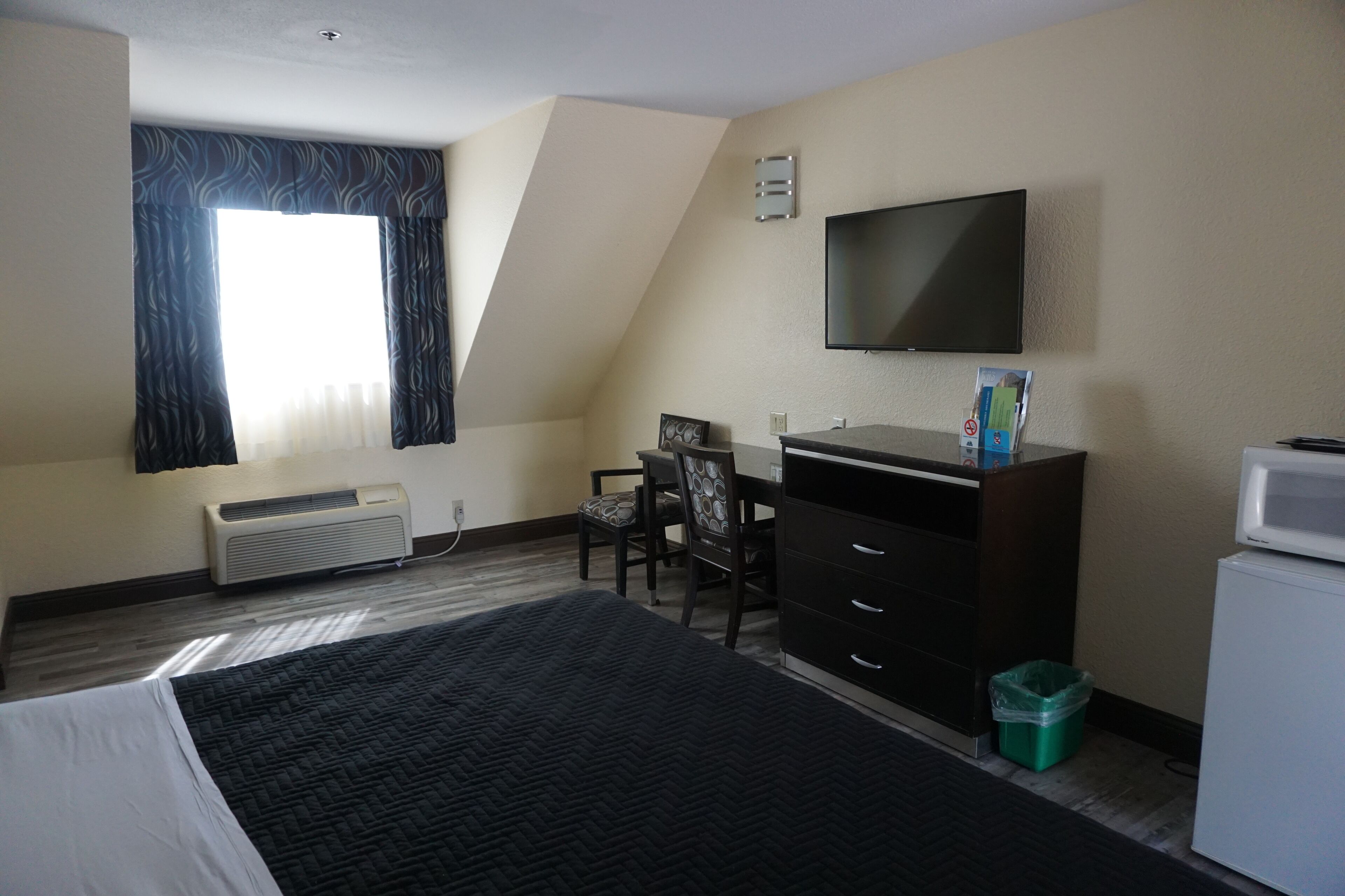 A hotel room with a bed, a desk with a chair, a television, a small refrigerator, and a window with curtains.