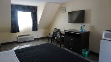 A hotel room with a bed, a desk with a chair, a television, a small refrigerator, and a window with curtains.
